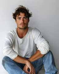 Wall Mural - A 30-year-old male model seated on the floor with legs stretched out and one arm resting on his knee, dressed in a fitted long-sleeve shirt and jeans. The white background enhances the casual yet thou