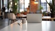 © Yi - Sleek modern office desk with an open laptop and coffee mug, set against a blurred background of a busy co-working space, representing productivity and contemporary work environments.