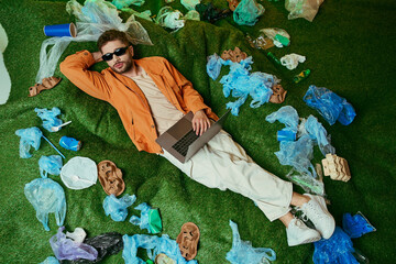 Wall Mural - A man lies on artificial grass surrounded by plastic waste, using a laptop.