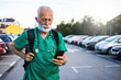 © Jelena Stanojkovic - Shot of a mature male nurse standing in a parking lot in his scrubs, while typing on his smartphone and smiling.