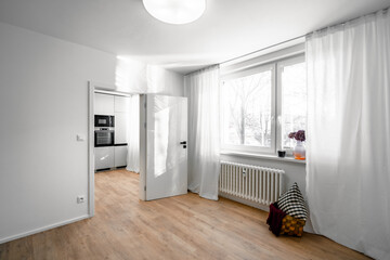 Wall Mural - A bright room with wooden parquet flooring, white walls, and a large window with white curtains. An open door reveals a modern kitchen in the background.