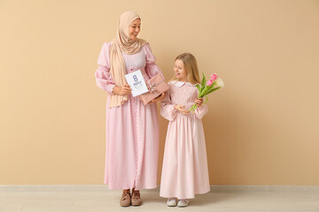 Canvas Print - Beautiful young Muslim woman holding gift box and her cute little daughter with bouquet of tulips near beige wall