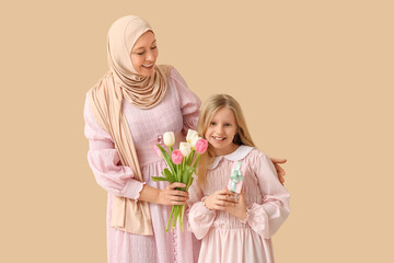 Poster - Beautiful young Muslim woman holding bouquet of tulips and her cute little daughter with gift box on beige background