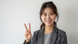 © Muhammad - Japanese woman casual business attire smiling making peace sign her right hand against white background She wearing gray blazer over striped shirt professional photography style left side of screen