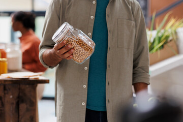 Wall Mural - Close-up of male customer carrying a jar of lentils while approaching checkout counter of local market. Selective focus on person grasping plastic free sustainable food product.