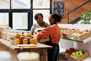 Wall Mural - Black woman wearing an apron is carrying boxes of fresh pesticide free produce from local farmer in eco friendly store. Female supplier delivers crates of farm grown products to bio food shop owner.