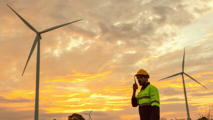 Wall Mural - man in a yellow and black safety vest is standing in front of three wind turbines. the sky is orange