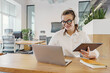 © muse studio - A young woman with headphones, glasses, and a tablet smiles while working on her laptop in a bright, modern office.