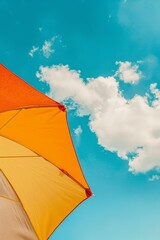 A vibrant orange and yellow beach umbrella stands proudly against a backdrop of a bright blue sky dotted with fluffy white clouds.