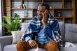 © Liubomir - Middle-aged man feeling unwell and covering himself with a blanket while sitting on a couch. He appears to have a cold, flu, and runny nose. The setting is a comfortable living room.