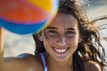 Wall Mural - A happy girl with dark hair and freckles, smiling while holding a colorful beach ball on a sunny beach, encapsulating youthful exuberance and playfulness in a lively setting.