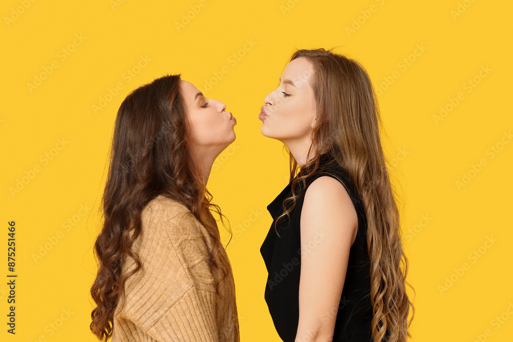 Beautiful young sisters kissing on yellow background