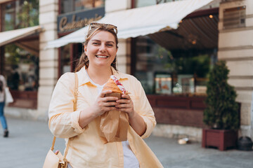 Beautiful overweight brunette 30s girl posing on the street, she has big croissant meal in hands. Unhealthy fattening food,high-calorie snack, eating on the go, take-out meals.