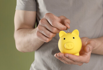 Poster - Man putting coin into yellow piggy bank on olive background, closeup