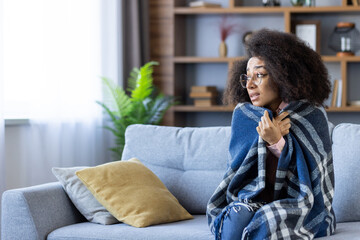 Woman sitting on a sofa wrapped in a warm blanket, visibly feeling cold. The indoor setting is cozy with decorative elements. The image portrays winter, warmth, comfort, and relaxation