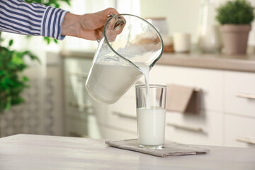 Wall Mural - Woman pouring fresh milk from jug into glass at light wooden table in kitchen, closeup