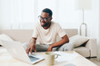 © SHOTPRIME STUDIO - Young African American freelancer working on a laptop while sitting on a modern sofa in his cozy home office With a smile on his face, he is immersed in his work, typing away on the keyboard The room