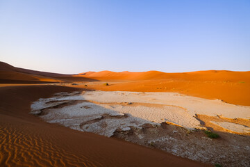 tunning landscape of Sossusvlei in the Namib Desert, Namibia. Orange sand dunes contrast with the white, cracked clay pan under a clear blue sky.