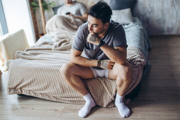 Wall Mural - Caucasian gay couple having argument with each other in bedroom. Frustrated gay man sitting on bed with pondering and sad look while another offended guy is sitting behind - Homosexual love concept