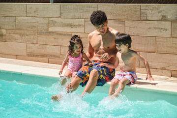 A man and his two children splashing water with their feet in a pool. Scene is happy and playful