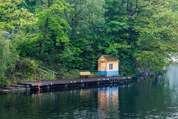 Wall Mural - Small empty wooden pier with a bench tool house on shore of Lake Robertville, green leaf trees in blurred background, cloudy day in Waimes, Belgium