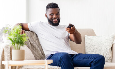 Wall Mural - Black man is sitting on a couch in a living room, wearing a white t-shirt and jeans. He is holding a remote control and watching television, with a plant on the table next to him.