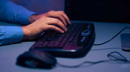 Poster - Cropped of a persons hands typing on a black keyboard. The image is taken at night, and the light from the computer screen illuminates the keyboard and the persons hands