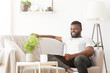 © Prostock-studio - Black man in a white t-shirt and black pants sits relaxed on a cream-colored couch in a brightly lit living room. The room is decorated with a potted plant and a floor lamp