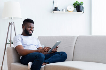 Wall Mural - Black man is sitting on a light gray sofa in a living room, looking at a tablet. He is smiling and wearing a white t-shirt and blue jeans. There is a floor lamp and a plant in the background.