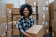 © Silga - Smiling warehouse worker holding a cardboard box, standing in a storage area with lots of stacked boxes shipping and logistics concept. Woman African American with curly hair holding box