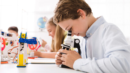Aufkleber - A young boy, wearing a white shirt, looks intently through a microscope, focusing on a slide. He is sitting at a desk in a classroom setting, surrounded by other students and educational materials