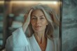 © Jane_S - Mature woman in a white robe, touching her hair, looking confidently into the mirror in a softly lit bathroom.