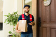 © AntonioDiaz - Latin delivery man with a cheerful smile delivering food to a house with a smartphone and paper bag