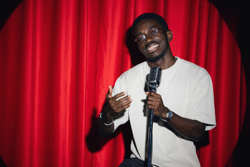 Wall Mural - Happy young african american man comic performing standup monologue on stage with red curtains. Concept comedian show open mike