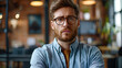 © Hoody Baba - Serious Young Man with Glasses and a Beard Looking at Camera in a Busy Office Environment