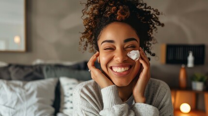 A woman applies face cream while sitting on her bed. Her skin care routine makes her happy.