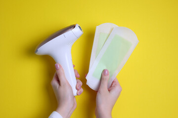 Wall Mural - A modern epilator for removing unwanted body hair at home and wax strips on a colored background in female hands