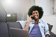 © JorSons/peopleimages.com - African woman, smile and phone call on sofa in house for conversation, networking and relax with laptop. Tech, talk and remote work as social media manager for company, agency or business in lounge