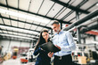 © Halfpoint - Two project managers standing in modern industrial factory, looking document in clipboard. Manufacturing facility with robotics, robotic arms and automation. Storing products and materials in