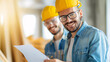 © eaglesky - Smiling construction workers wearing yellow hard hats and glasses, reviewing blueprints in a bright building site.