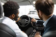 © sofiko14 - Salesman pointing at dashboard while client holding steering wheel, interaction during car purchase . Caucasian car dealer sit inside vehicle with African American buyer discussing car features.