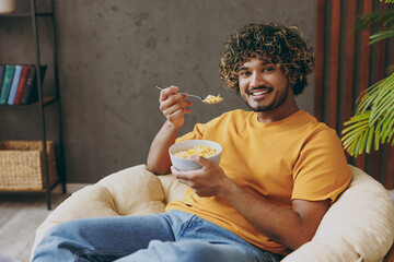 Wall Mural - Young Indian man wearing orange casual clothes eat breakfast muesli cereals with milk fruit in bowl sits in armchair stay at home hotel flat rest relax spend free time in living room. Lounge concept.