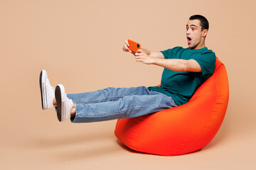 Sticker - Young gambling middle eastern man in blue t-shirt casual clothes sit in bag chair use play racing app on mobile cell phone hold gadget smartphone for pc video games isolated on plain beige background.