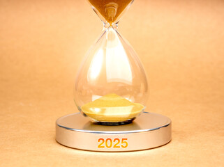 Wall Mural - Coming soon to new year 2025, business and technology concept. Close-up hourglass with gold sand inside, on round silver chrome podium with 2025 year calendar number on brown background, minimalist.