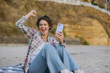 Wall Mural - Young happy cool woman wear shirt casual clothes hold in hand use mobile cell phone do winner gesture sit rest on sea ocean sand shore beach outdoor seaside in summer day free time. Lifestyle concept.