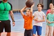 © LIGHTFIELD STUDIOS - A group of young children standing around a basketball as their teacher instructs them in a bright, lively classroom setting.