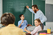 © LIGHTFIELD STUDIOS - A group of diverse children listening attentively to their male teachers instruction at a desk in front of a blackboard in a bright and lively classroom setting.