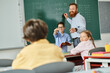 © LIGHTFIELD STUDIOS - A male teacher stands confidently in front of a blackboard, instructing a group of children in a bright and lively classroom setting.