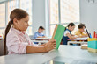 © LIGHTFIELD STUDIOS - A young girl sits at a table, engrossed in a book in a bright classroom setting.