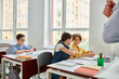 © LIGHTFIELD STUDIOS - A group of children sit at desks in a bright classroom, focusing on a man teacher giving instructions.